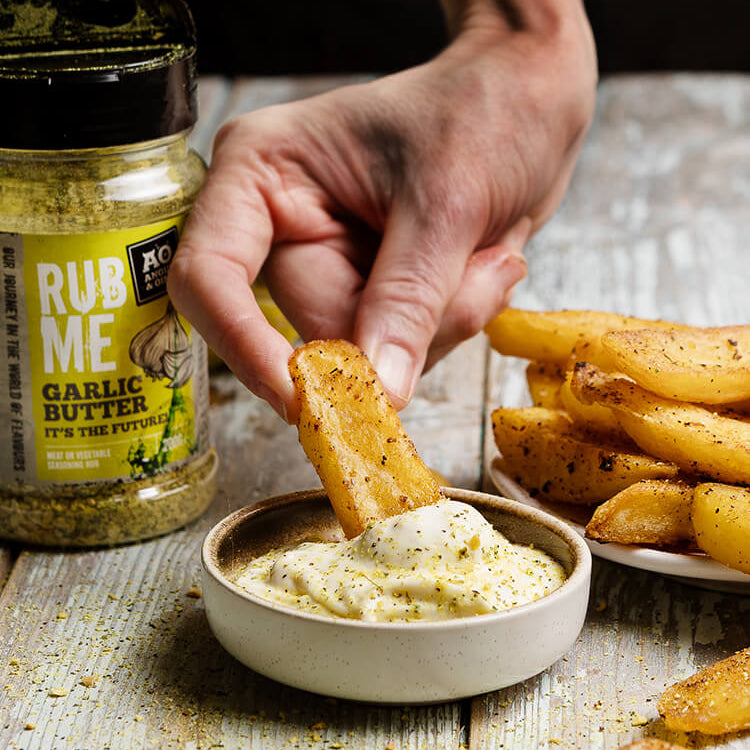 Person dipping a potato wedge into garlic butter with 'Rub Me Garlic Butter' seasoning jar on a wooden table.