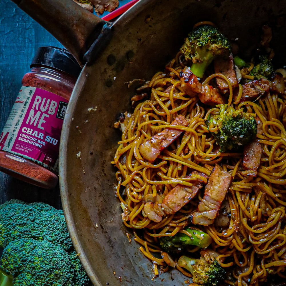 Frying pan with noodles, broccoli, and pork on a blue background with a jar of angus & oink char siu seasoning.