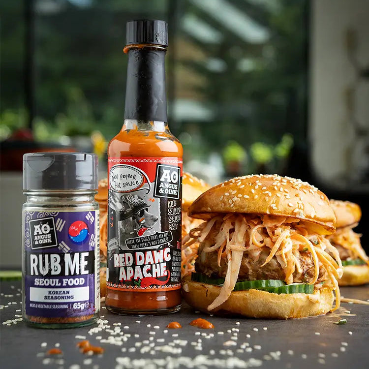 Bottle of 'Red Dawg Apache' sauce and 'Rub Me  Korean' seasoning with a burger on a dark surface.